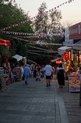 A lively outdoor market scene with people walking on a paved walkway, some pushing strollers. Stalls filled with various goods line the sides, and colorful flags are strung above. Trees provide a backdrop, and some neon signs illuminate the area.