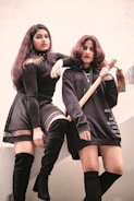 Two individuals are posing confidently. One person is standing on a ledge, wearing a black outfit with a skirt and thigh-high boots. The other is holding a tool, dressed in a long dark top and knee-high socks. Their expressions are serious and intense, suggesting a mood of defiance or empowerment.