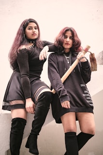 Two individuals are posing confidently. One person is standing on a ledge, wearing a black outfit with a skirt and thigh-high boots. The other is holding a tool, dressed in a long dark top and knee-high socks. Their expressions are serious and intense, suggesting a mood of defiance or empowerment.