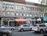 A city street scene with a brick building featuring multiple businesses. The storefronts include a café, a quirky restaurant with a colorful awning, and a burrito shop. Cars are parked along the street, and a few are driving by.