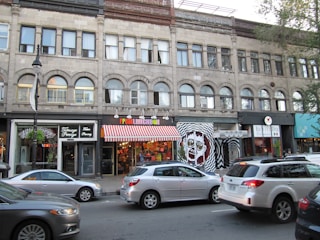 A city street scene with a brick building featuring multiple businesses. The storefronts include a café, a quirky restaurant with a colorful awning, and a burrito shop. Cars are parked along the street, and a few are driving by.