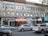 A city street scene with a brick building featuring multiple businesses. The storefronts include a café, a quirky restaurant with a colorful awning, and a burrito shop. Cars are parked along the street, and a few are driving by.