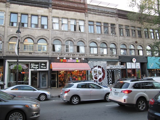 A city street scene with a brick building featuring multiple businesses. The storefronts include a caf&eacute;, a quirky restaurant with a colorful awning, and a burrito shop. Cars are parked along the street, and a few are driving by.