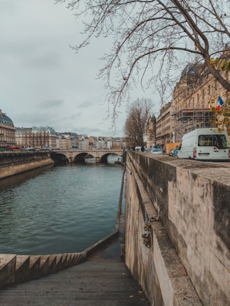 A scenic view of a river with a stone bridge in the background. Bare tree branches hang over a cobblestone pathway that leads down to the water. On the right side, there are classical buildings lining the street and a white van parked nearby. A French flag is visible on a building.