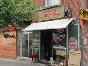 A quaint storefront with a brick exterior and an awning that reads 'Conserve Montreal'. The entrance is glass with a sign indicating 'Groceries'. A small stool and what appears to be a statue of a dog are placed outside. There is a neon 'Ouvert' sign visible in the window, and various product advertisements and boards are displayed on the exterior walls.