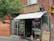 A quaint storefront with a brick exterior and an awning that reads 'Conserve Montreal'. The entrance is glass with a sign indicating 'Groceries'. A small stool and what appears to be a statue of a dog are placed outside. There is a neon 'Ouvert' sign visible in the window, and various product advertisements and boards are displayed on the exterior walls.