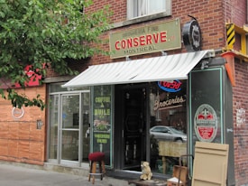 A quaint storefront with a brick exterior and an awning that reads 'Conserve Montreal'. The entrance is glass with a sign indicating 'Groceries'. A small stool and what appears to be a statue of a dog are placed outside. There is a neon 'Ouvert' sign visible in the window, and various product advertisements and boards are displayed on the exterior walls.