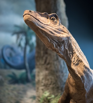 A detailed and lifelike dinosaur model is depicted, specifically showing the head and neck. The background includes blurred elements such as trees and plants, suggestive of a prehistoric environment.