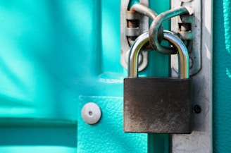 A close-up of a sturdy silver padlock securing a bright turquoise-painted door with a thick, rusted chain. The metal lock has a reflective surface, and the bolt is fastened tightly to ensure security.