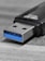 A close-up view of a USB flash drive on a textured surface, highlighting the metal connector with a blue plastic insert characteristic of USB 3.0. The drive's black housing is partially visible.