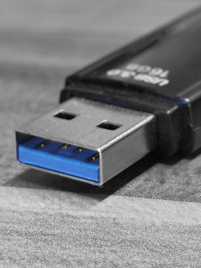 A close-up view of a USB flash drive on a textured surface, highlighting the metal connector with a blue plastic insert characteristic of USB 3.0. The drive's black housing is partially visible.