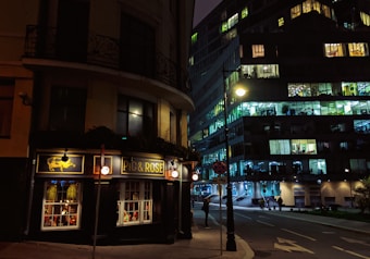 A dimly lit street corner featuring a pub named 'Pig & Rose' with a dark, elegant exterior. The street is flanked by a modern office building with numerous illuminated windows suggesting a bustling environment inside. Streetlights cast a warm glow over the scene, and a few people are walking along the sidewalk.