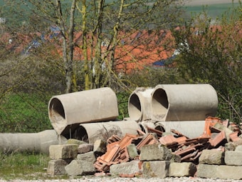 A pile of large concrete pipes and bricks lies in a grassy area with trees in the foreground. The background features a view of a village with red-roofed houses and fields extending into the distance. Sunlight enhances the natural colors and creates a rural and somewhat abandoned atmosphere.