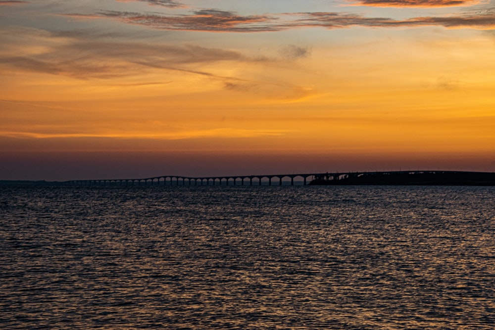 the sun is setting over the ocean with a bridge in the distance