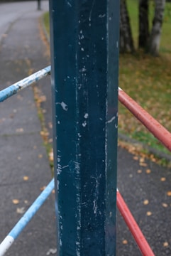 A close-up view of a weathered metal barrier with faded paint showing signs of wear and tear. The pole is primarily blue, and the railings are painted in blue, white, and red. The background reveals a path with fallen leaves scattered on the ground and a grassy area with some trees.
