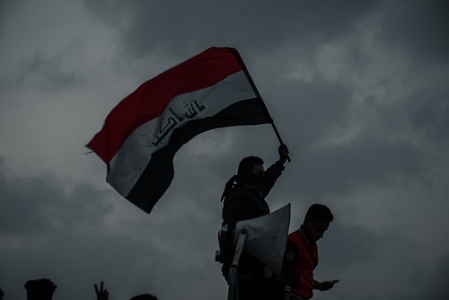 A person is holding a large, waving flag with Arabic script against a dramatic, cloudy sky. The silhouette suggests a sense of defiance or protest, with another person nearby holding a phone.