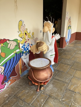A character statue in a white robe and red cape stands in front of a large pot, holding an object in its hands. The scene is set against a backdrop of colorful cartoon characters on a wall.