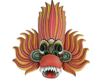 A vibrant and intricately designed mask with exaggerated features, including large eyes, sharp teeth, and flowing, flame-like elements emanating from the sides and top. The colors are vivid, with a mix of reds, yellows, and greens, giving it a dynamic and striking appearance.