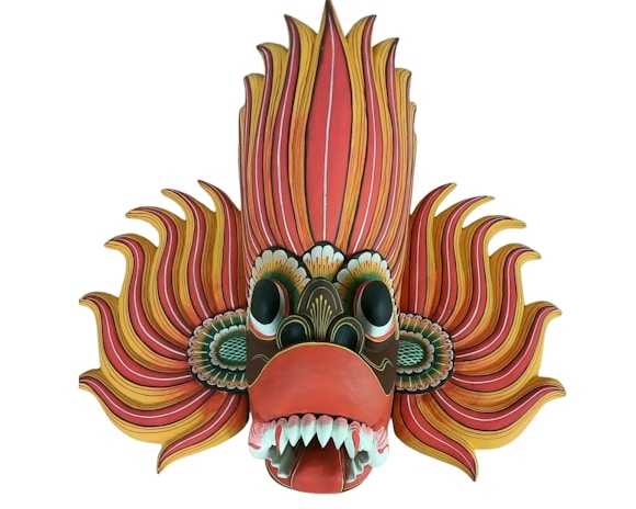 A vibrant and intricately designed mask with exaggerated features, including large eyes, sharp teeth, and flowing, flame-like elements emanating from the sides and top. The colors are vivid, with a mix of reds, yellows, and greens, giving it a dynamic and striking appearance.
