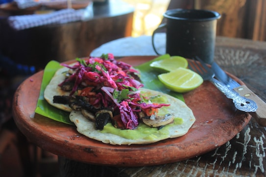 A rustic wooden table holds a clay plate with a taco topped with vibrant red cabbage, mushrooms, and greens on a bed of guacamole. A lime cut in halves sits beside it, along with a fork and knife. A dark-colored mug is in the background.