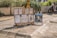 Several portrait sketches and a painted caricature placed on an easel stand on a sunny paved street. The sketches depict different individuals, some in pairs and others singularly. There is a scooter parked nearby and green plants in large pots adding a touch of nature to the urban setting.