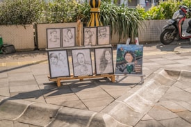 Several portrait sketches and a painted caricature placed on an easel stand on a sunny paved street. The sketches depict different individuals, some in pairs and others singularly. There is a scooter parked nearby and green plants in large pots adding a touch of nature to the urban setting.