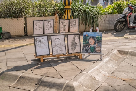 Several portrait sketches and a painted caricature placed on an easel stand on a sunny paved street. The sketches depict different individuals, some in pairs and others singularly. There is a scooter parked nearby and green plants in large pots adding a touch of nature to the urban setting.