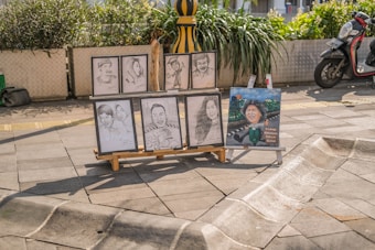 Several portrait sketches and a painted caricature placed on an easel stand on a sunny paved street. The sketches depict different individuals, some in pairs and others singularly. There is a scooter parked nearby and green plants in large pots adding a touch of nature to the urban setting.