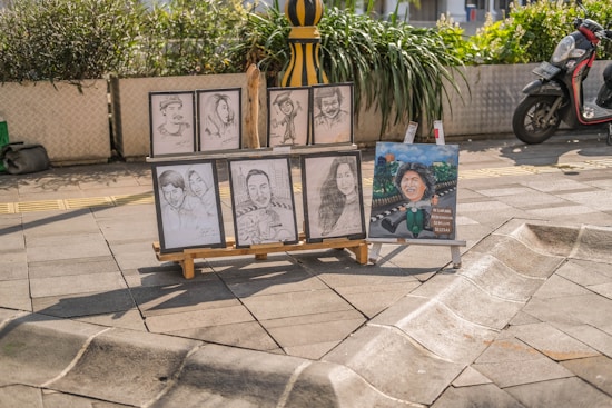 Several portrait sketches and a painted caricature placed on an easel stand on a sunny paved street. The sketches depict different individuals, some in pairs and others singularly. There is a scooter parked nearby and green plants in large pots adding a touch of nature to the urban setting.