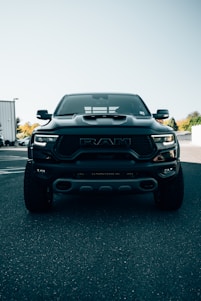 A large, black pickup truck is parked on an asphalt surface under a clear sky. The truck's front grille is prominently displayed, with the logo clearly visible. The background includes partial views of buildings and trees, adding an urban touch.