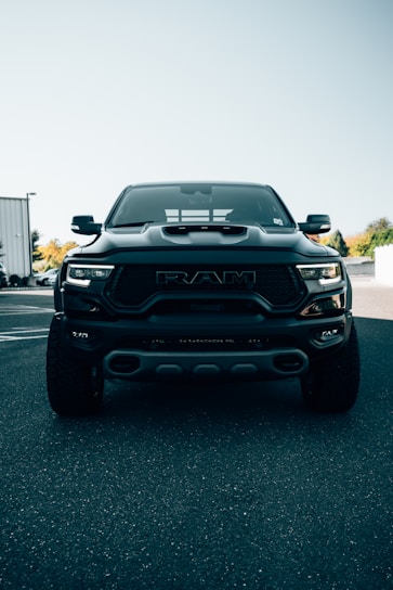 A large, black pickup truck is parked on an asphalt surface under a clear sky. The truck's front grille is prominently displayed, with the logo clearly visible. The background includes partial views of buildings and trees, adding an urban touch.