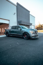 A sleek, modern pickup truck with a metallic finish is parked in front of a large industrial building. The building has a white and gray facade with an open garage door on the left. The truck is detailed with black wheels and red calipers, giving it a sporty appearance. The background includes a clear sky and some greenery.