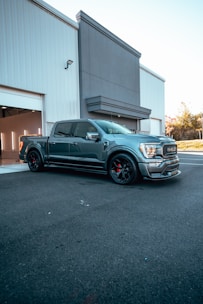 A sleek, modern pickup truck with a metallic finish is parked in front of a large industrial building. The building has a white and gray facade with an open garage door on the left. The truck is detailed with black wheels and red calipers, giving it a sporty appearance. The background includes a clear sky and some greenery.