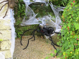 A large, artificial black spider with long legs is set against a background of stone and foliage. It sits amidst white artificial spider webs, surrounded by green leaves and vibrant red flowers. The scene appears to be part of a Halloween decoration.