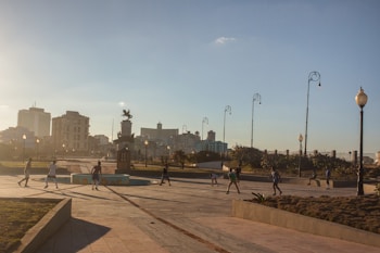 A cityscape features a public square or open space with a group of people engaged in various activities under street lamps. There is a prominent statue situated in the middle, surrounded by well-kept pathways and manicured shrubs. In the background, several buildings and skyscrapers are visible against a clear sky, suggesting an urban setting.