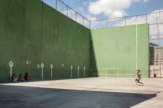 A young person is playing tennis against a large green wall, likely a handball or racquetball court, while three people sit in the shade nearby observing. The court is enclosed with a wire fence on top, and a small football goal is painted on the wall. The ground is concrete and part of it is in shadow.