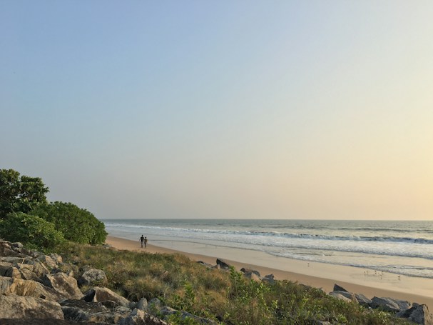 A serene beach landscape with gentle waves meeting the shore under a clear sky. Along the coast, a couple is walking, surrounded by lush greenery and scattered rocks in the foreground. The ocean stretches towards the horizon, reflecting the soft sunlight.
