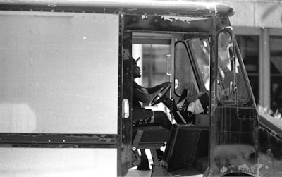 A black and white photograph of a person driving a delivery or utility truck. The person is sitting in the driver's seat, holding the steering wheel, and wearing a cap. The truck's interior and partially visible exterior indicate wear, suggesting heavy use.