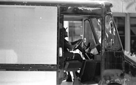 A black and white photograph of a person driving a delivery or utility truck. The person is sitting in the driver's seat, holding the steering wheel, and wearing a cap. The truck's interior and partially visible exterior indicate wear, suggesting heavy use.
