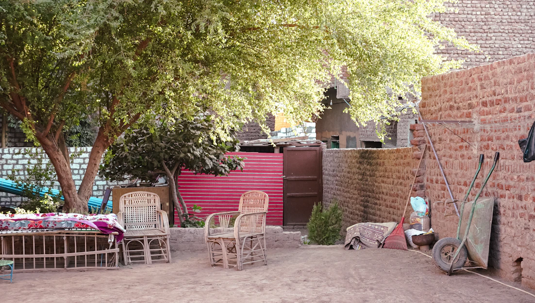 The image depicts an outdoor courtyard setting featuring a variety of objects and furniture. There are wicker chairs and a bench with colorful cushions, positioned under the shade of a large tree. Surrounding the area are brick walls and a door at the back, along with a vibrant red panel. A wheelbarrow and some stacked utility items are visible on the right side, adding to the rustic charm.