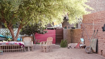 The image depicts an outdoor courtyard setting featuring a variety of objects and furniture. There are wicker chairs and a bench with colorful cushions, positioned under the shade of a large tree. Surrounding the area are brick walls and a door at the back, along with a vibrant red panel. A wheelbarrow and some stacked utility items are visible on the right side, adding to the rustic charm.