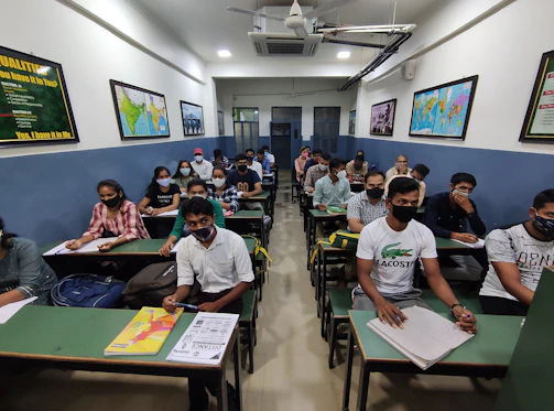 A classroom filled with students wearing masks, seated at green desks arranged in rows. The walls are adorned with maps and educational posters. The room is well-lit with overhead fluorescent lighting, and students appear to be taking notes or listening attentively.