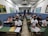 A classroom filled with students wearing masks, seated at green desks arranged in rows. The walls are adorned with maps and educational posters. The room is well-lit with overhead fluorescent lighting, and students appear to be taking notes or listening attentively.