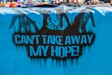 A vibrant, blue cloth features black graffiti art, including silhouetted figures resembling hands breaking free from chains. The bold, black text on the cloth reads 'CAN'T TAKE AWAY MY HOPE!' The top of the image displays a variety of colorful, beaded bracelets and necklaces.