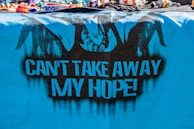 A vibrant, blue cloth features black graffiti art, including silhouetted figures resembling hands breaking free from chains. The bold, black text on the cloth reads 'CAN'T TAKE AWAY MY HOPE!' The top of the image displays a variety of colorful, beaded bracelets and necklaces.
