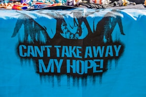 A vibrant, blue cloth features black graffiti art, including silhouetted figures resembling hands breaking free from chains. The bold, black text on the cloth reads 'CAN'T TAKE AWAY MY HOPE!' The top of the image displays a variety of colorful, beaded bracelets and necklaces.