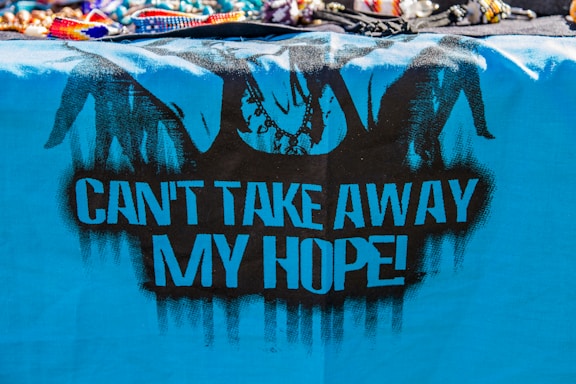 A vibrant, blue cloth features black graffiti art, including silhouetted figures resembling hands breaking free from chains. The bold, black text on the cloth reads 'CAN'T TAKE AWAY MY HOPE!' The top of the image displays a variety of colorful, beaded bracelets and necklaces.