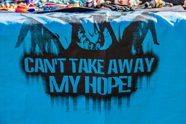 A vibrant, blue cloth features black graffiti art, including silhouetted figures resembling hands breaking free from chains. The bold, black text on the cloth reads 'CAN'T TAKE AWAY MY HOPE!' The top of the image displays a variety of colorful, beaded bracelets and necklaces.