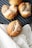 A set of freshly baked bread rolls are sprinkled with coarse salt. They are resting on a dark ridged baking tray, with one roll placed on a beige cloth beside the tray. The texture of the bread is golden brown with a crusty exterior, and the background features a soft fabric.
