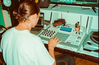 A person with long hair tied back operates music production equipment, including an AKAI MPC and a laptop, in a studio setting. Additional equipment like a turntable and speakers are visible, with various items such as a jar and pens placed on a desk.
